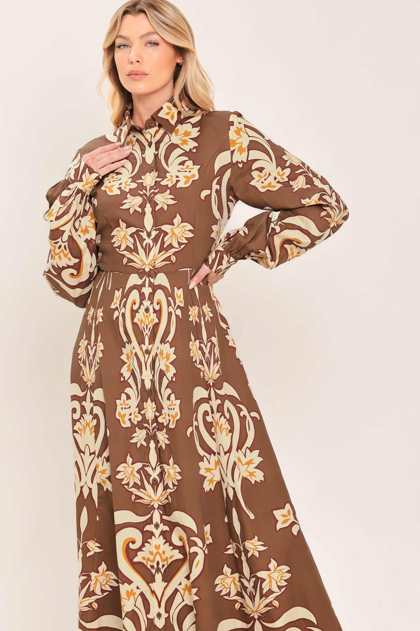 A woman wearing a long-sleeve, high-neck button up maxi dress with a brown base and a beige and gold baroque-style floral pattern | Flying Tomato