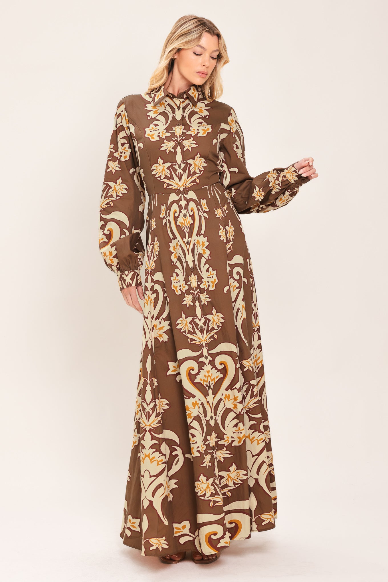 A woman wearing a long-sleeve, high-neck button up maxi dress with a brown base and a beige and gold baroque-style floral pattern | Flying Tomato