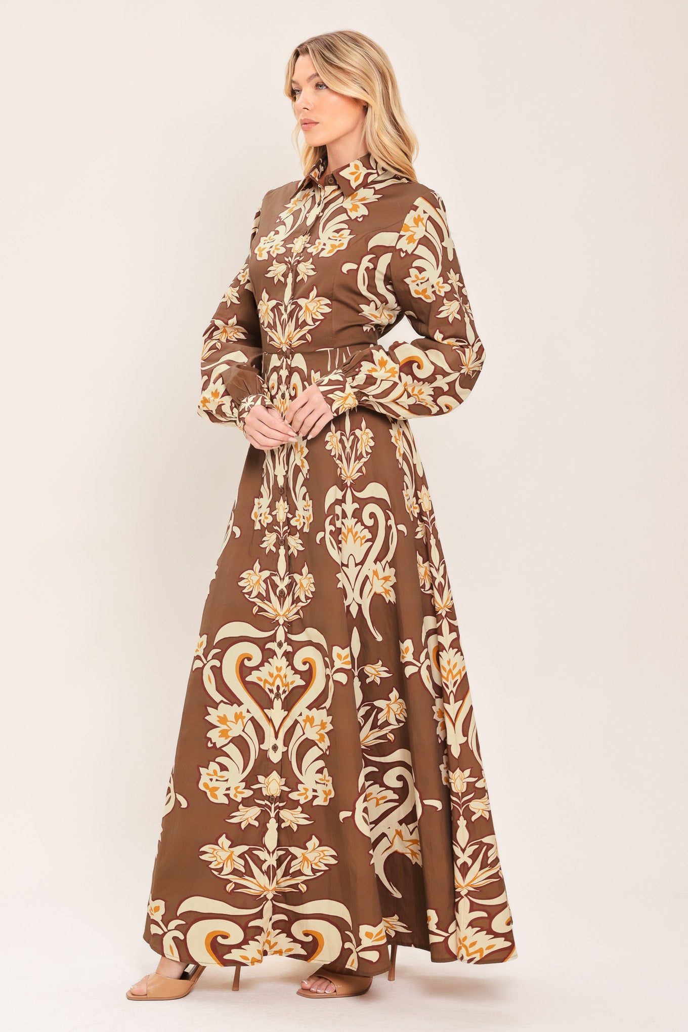 A woman wearing a long-sleeve, high-neck button up maxi dress with a brown base and a beige and gold baroque-style floral pattern | Flying Tomato