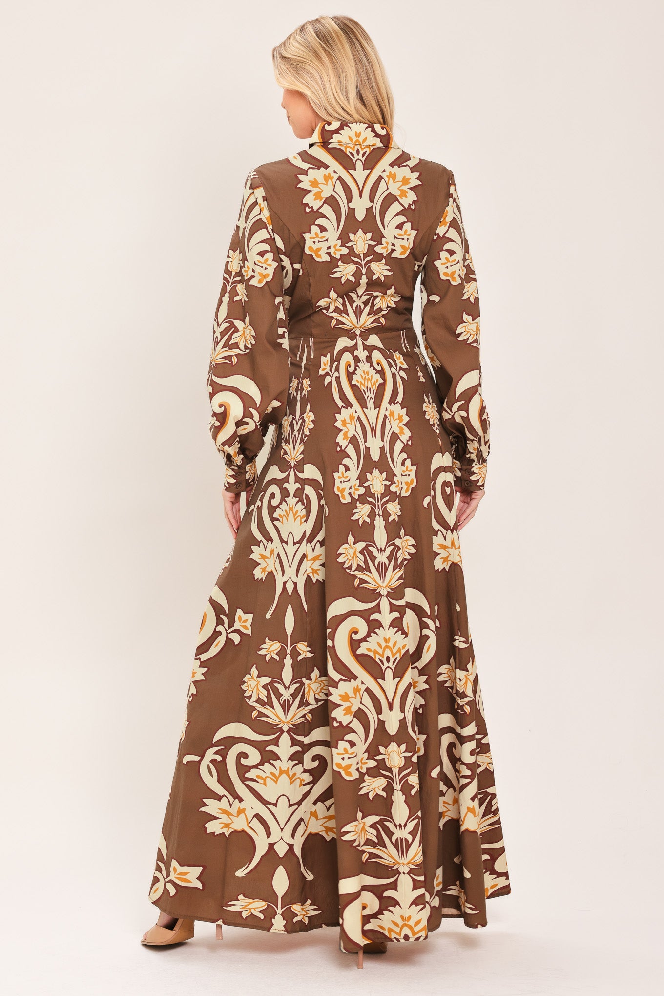 A woman wearing a long-sleeve, high-neck button up maxi dress with a brown base and a beige and gold baroque-style floral pattern | Flying Tomato