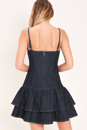 Woman wearing a dark blue denim mini dress featuring straight neckline straps, drop waist with double ruffle layers hem and back zipper closure | Flying Tomato