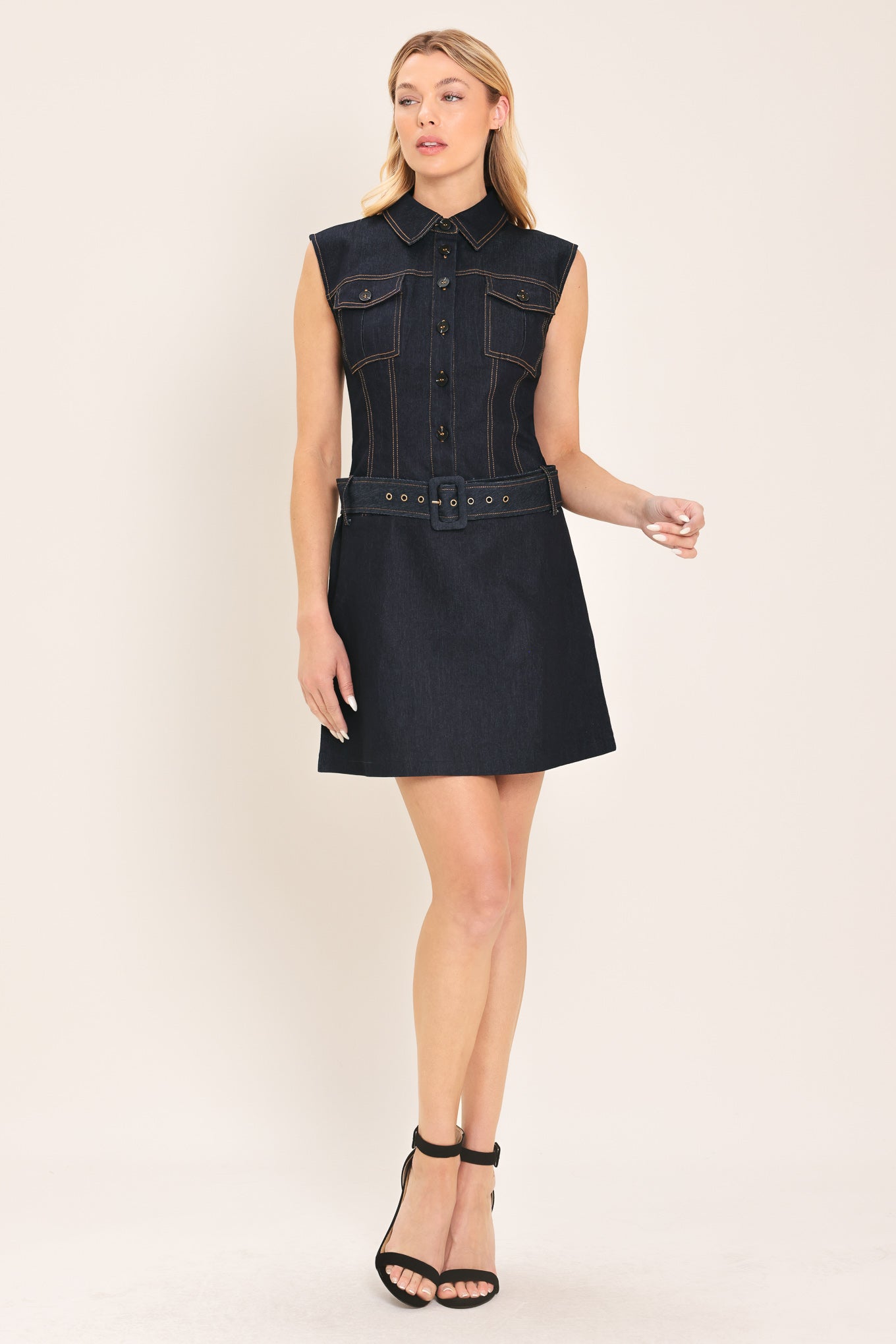 Woman wearing a sleeveless dark denim mini dress featuring collar, front button down and drop waist with belt | Flying Tomato