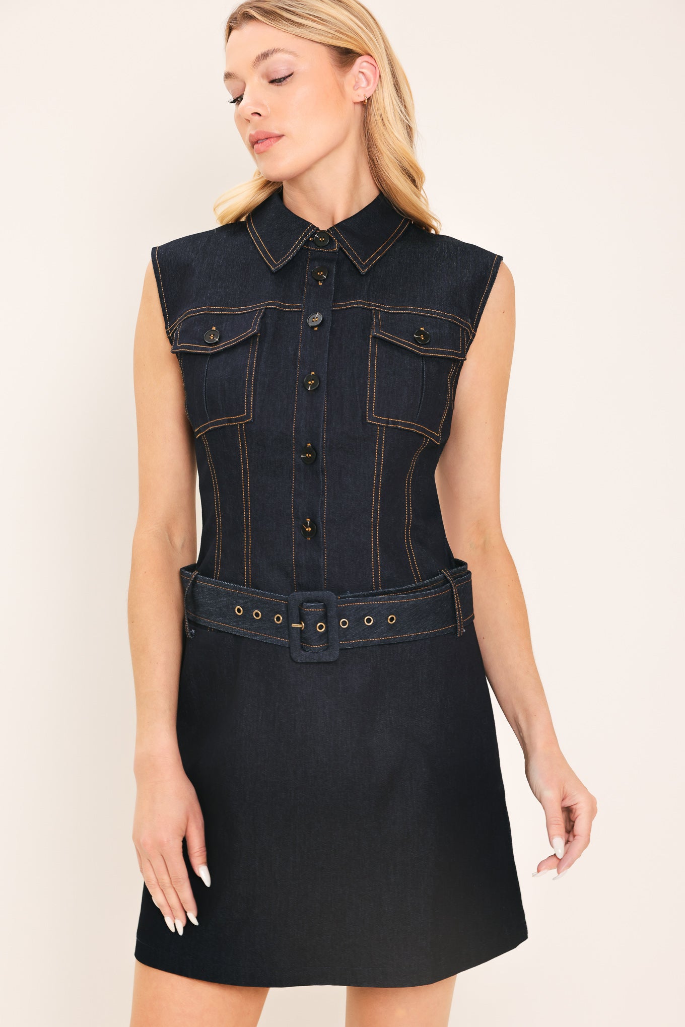 Woman wearing a sleeveless dark denim mini dress featuring collar, front button down and drop waist with belt | Flying Tomato