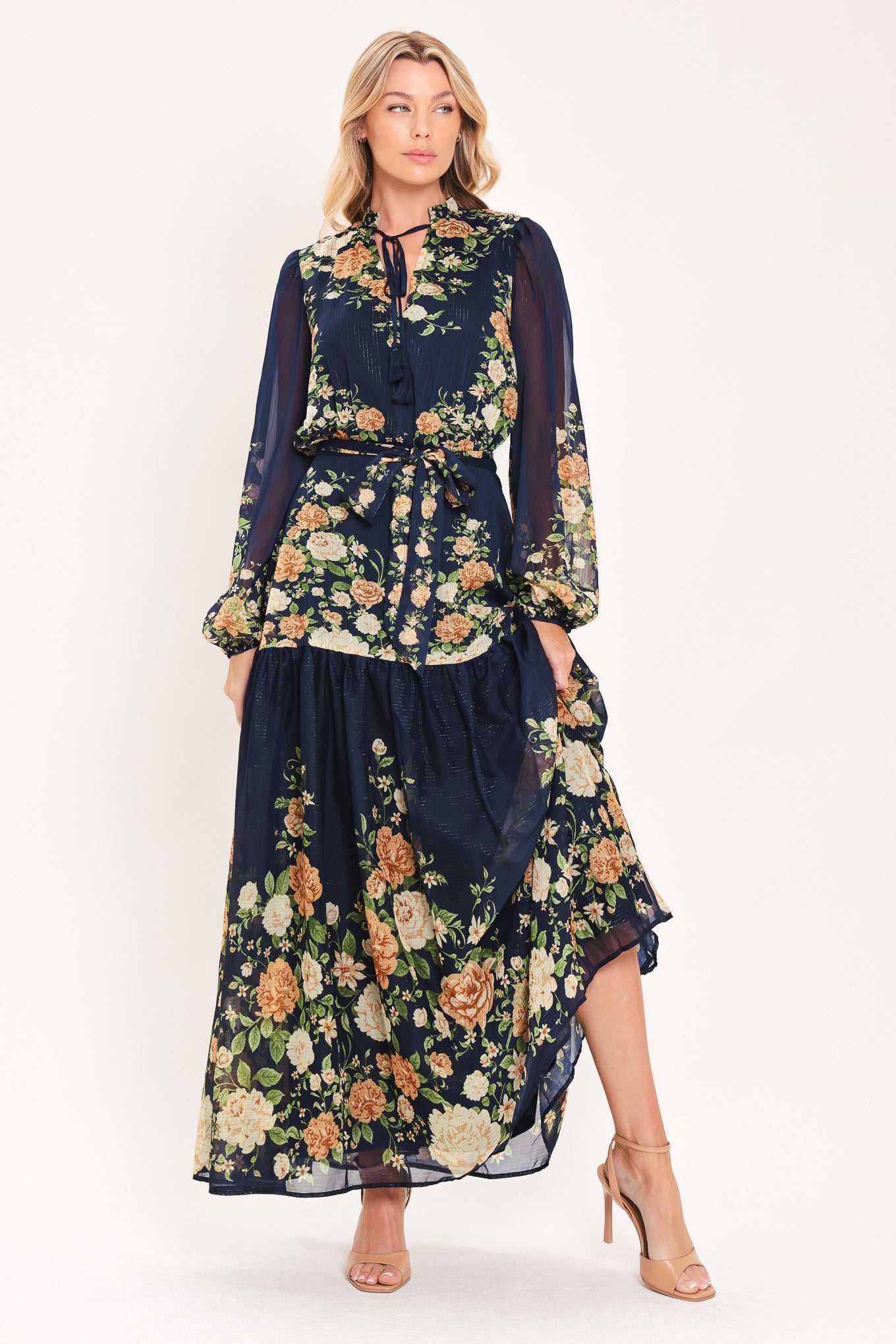 A woman modeling a navy blue long-sleeve maxi dress with a floral print. The dress features a tie-neck detail, a matching waist tie, and a tiered skirt | Flying Tomato
