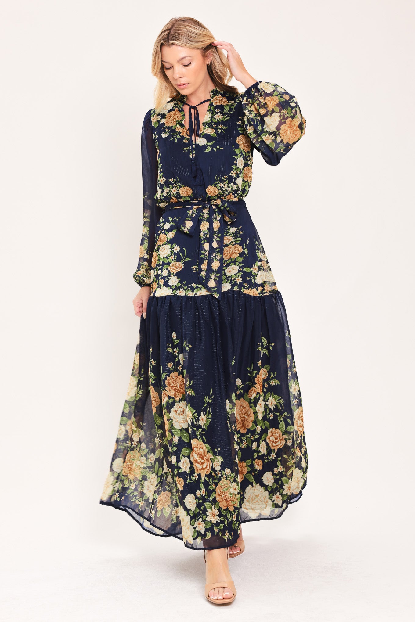 A woman modeling a navy blue long-sleeve maxi dress with a floral print. The dress features a tie-neck detail, a matching waist tie, and a tiered skirt | Flying Tomato