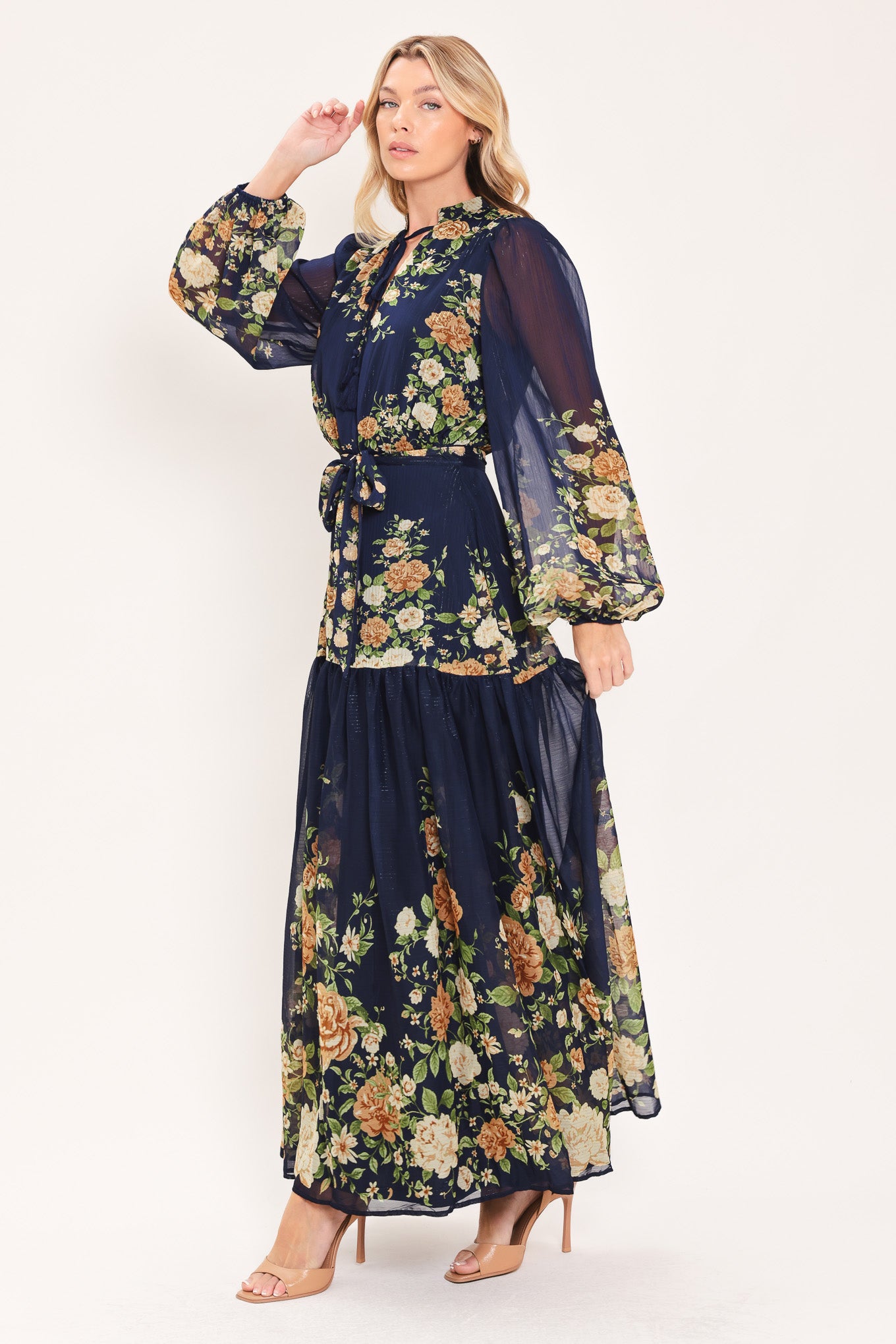 A woman modeling a navy blue long-sleeve maxi dress with a floral print. The dress features a tie-neck detail, a matching waist tie, and a tiered skirt | Flying Tomato