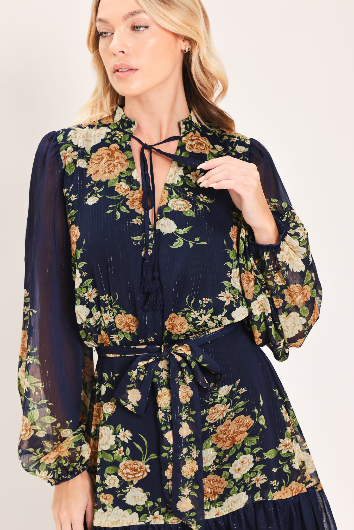 A woman modeling a navy blue long-sleeve maxi dress with a floral print. The dress features a tie-neck detail, a matching waist tie, and a tiered skirt | Flying Tomato