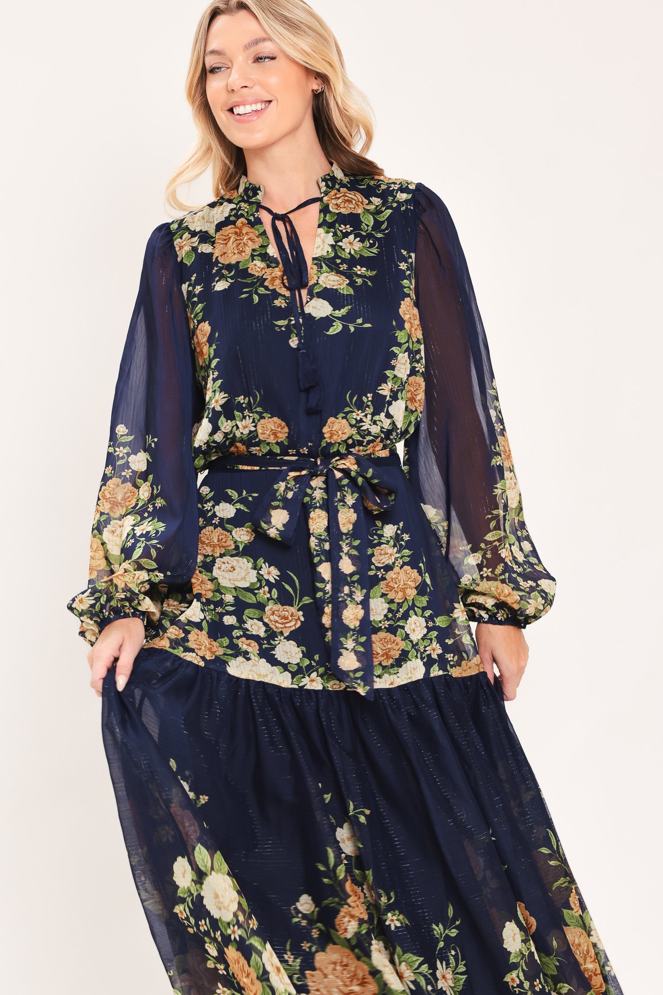 A woman modeling a navy blue long-sleeve maxi dress with a floral print. The dress features a tie-neck detail, a matching waist tie, and a tiered skirt | Flying Tomato