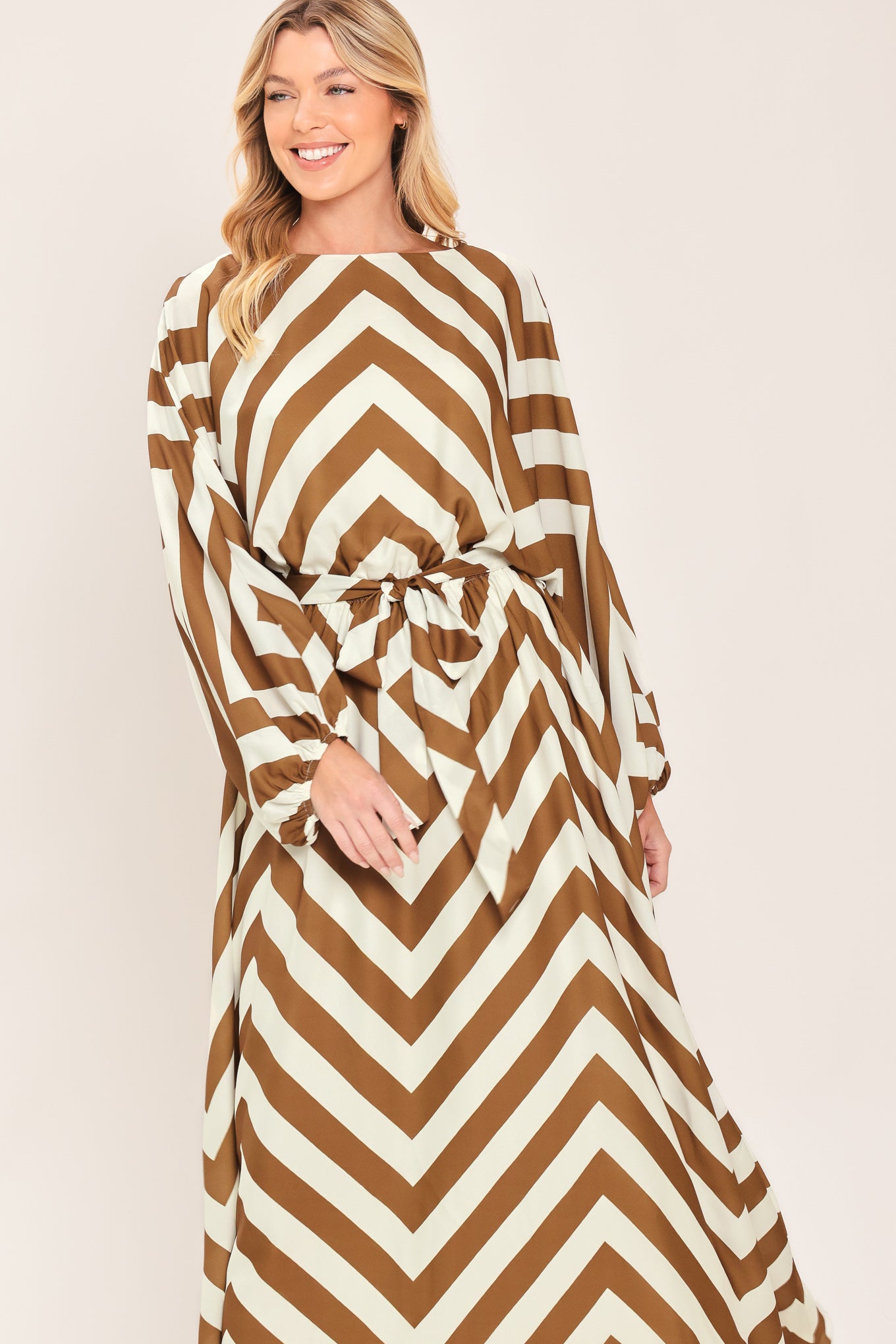 A woman modeling a long-sleeve maxi dress with a bold brown and ivory chevron pattern, featuring a tie waist belt | Flying Tomato