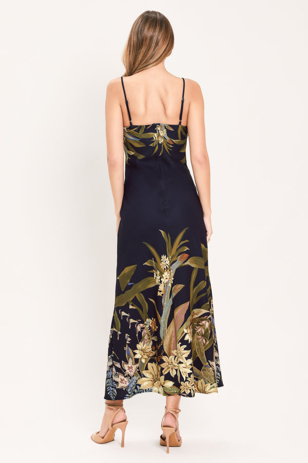 A woman wearing a sleeveless midi dress with a deep purple base and a tropical floral print in beige, gold, and muted green | Flying Tomato