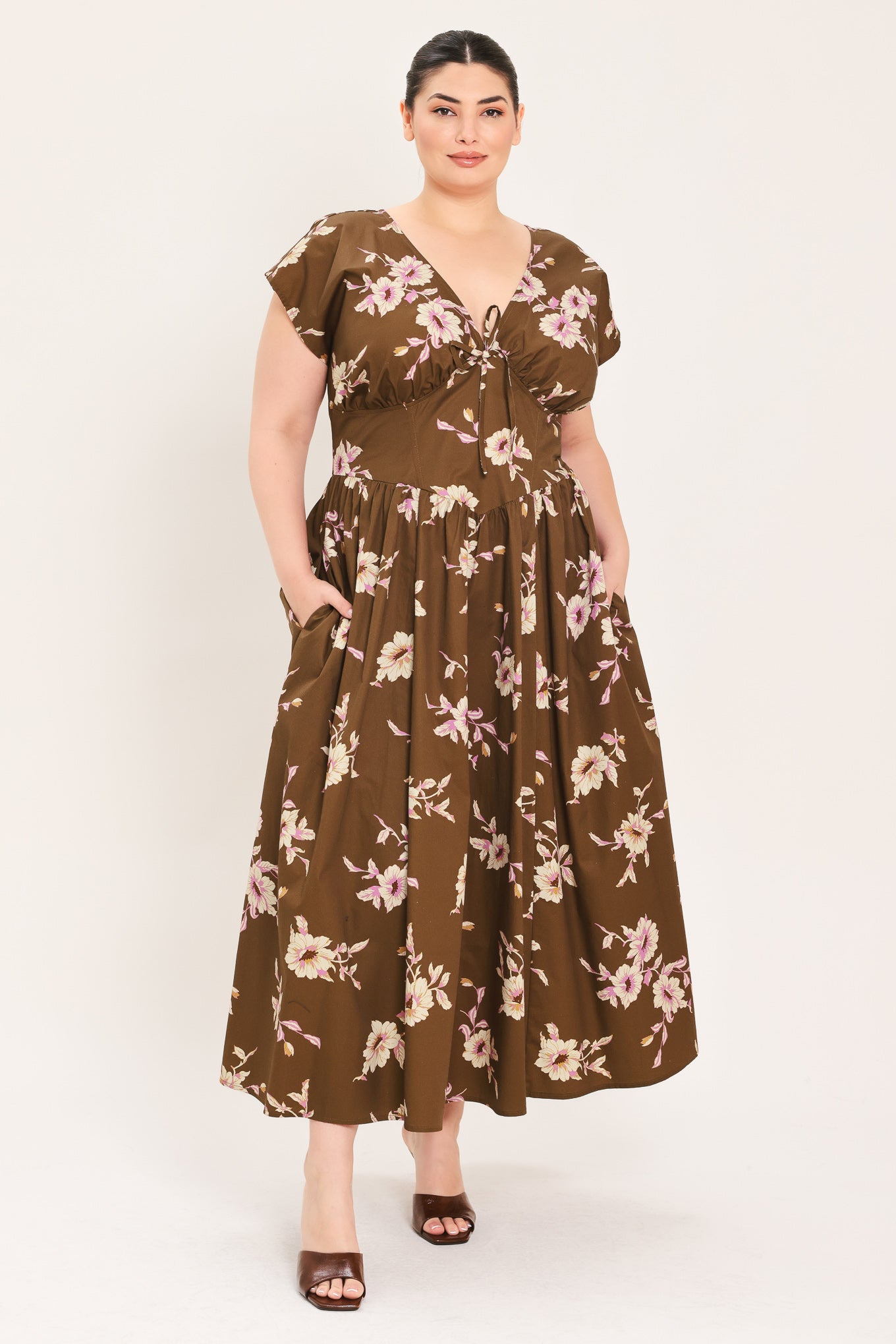 A woman modeling a brown midi dress with a floral print in pink and beige, featuring short puff sleeves, a V-neckline with a tie-front detail, and side pockets | Plus Curve Flying Tomato