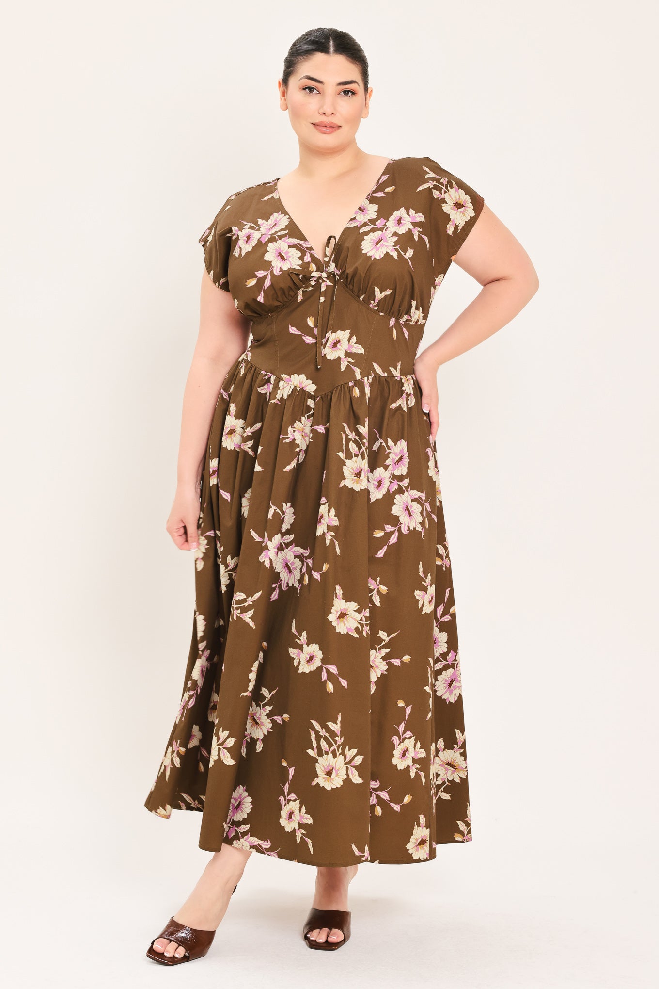A woman modeling a brown midi dress with a floral print in pink and beige, featuring short puff sleeves, a V-neckline with a tie-front detail, and side pockets | Plus Curve Flying Tomato