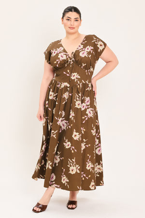 A woman modeling a brown midi dress with a floral print in pink and beige, featuring short puff sleeves, a V-neckline with a tie-front detail, and side pockets | Plus Curve Flying Tomato