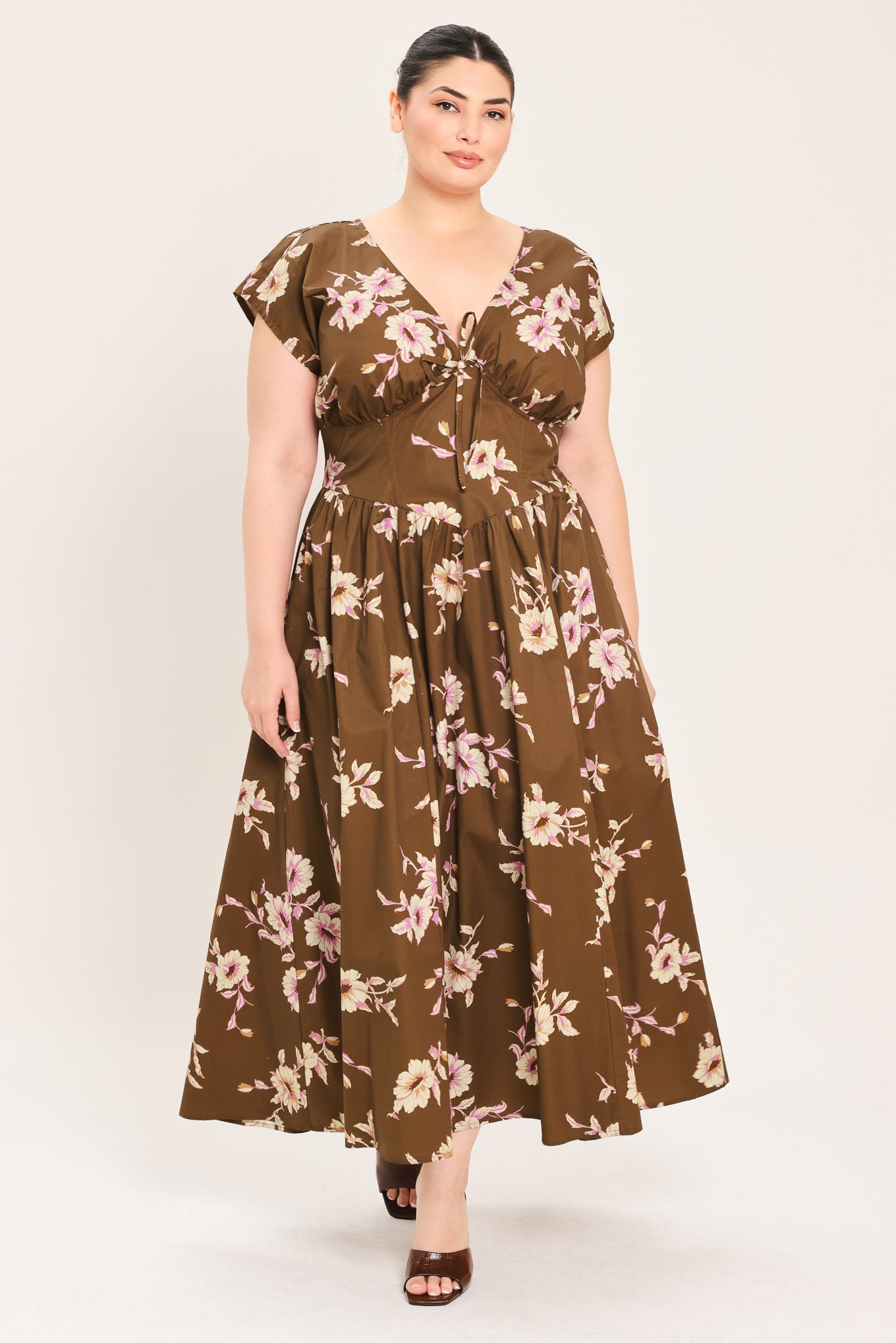 A woman modeling a brown midi dress with a floral print in pink and beige, featuring short puff sleeves, a V-neckline with a tie-front detail, and side pockets | Plus Curve Flying Tomato