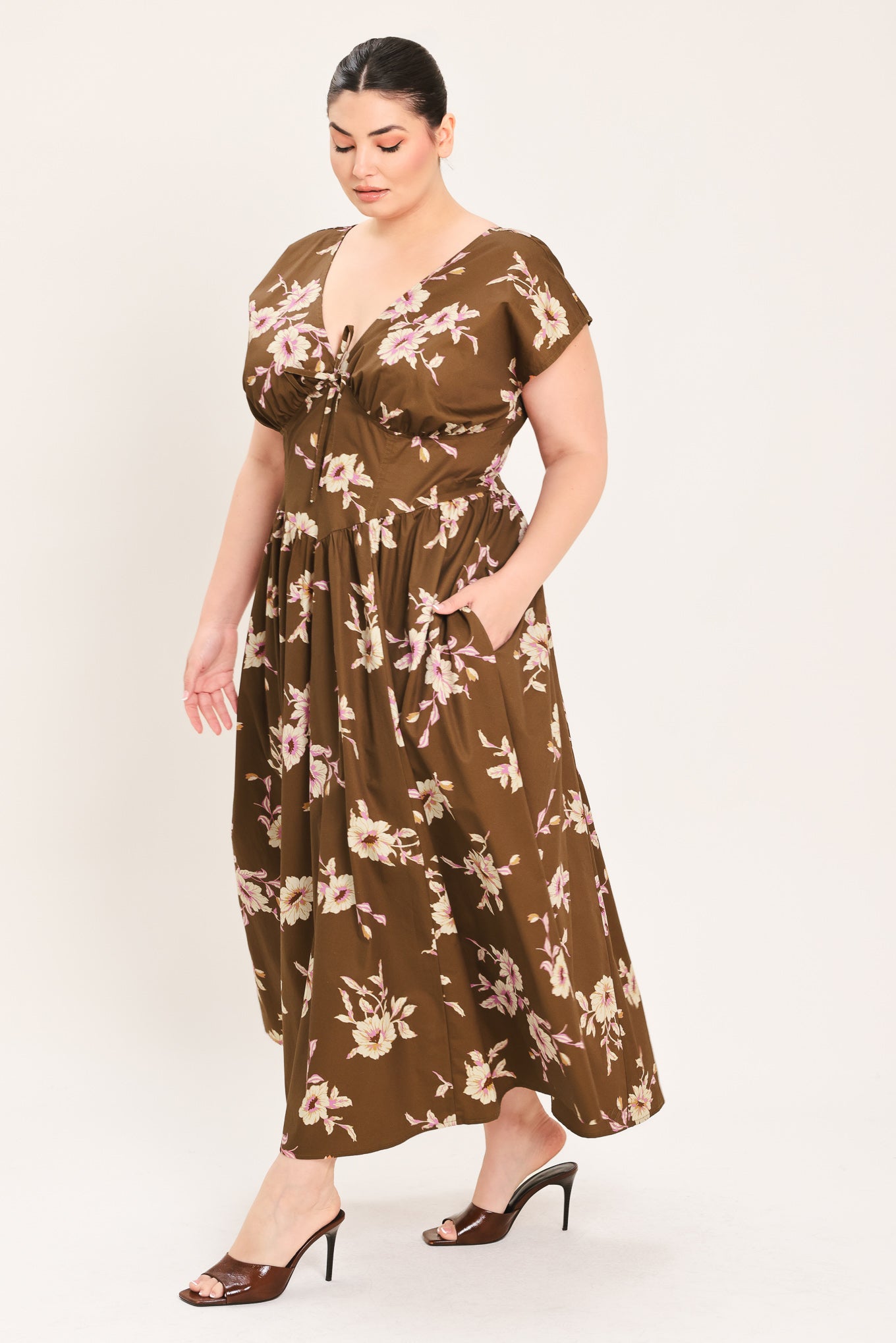 A woman modeling a brown midi dress with a floral print in pink and beige, featuring short puff sleeves, a V-neckline with a tie-front detail, and side pockets | Plus Curve Flying Tomato