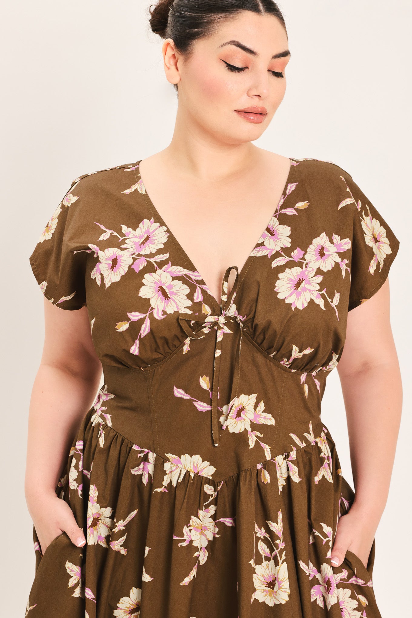 A woman modeling a brown midi dress with a floral print in pink and beige, featuring short puff sleeves, a V-neckline with a tie-front detail, and side pockets | Plus Curve Flying Tomato