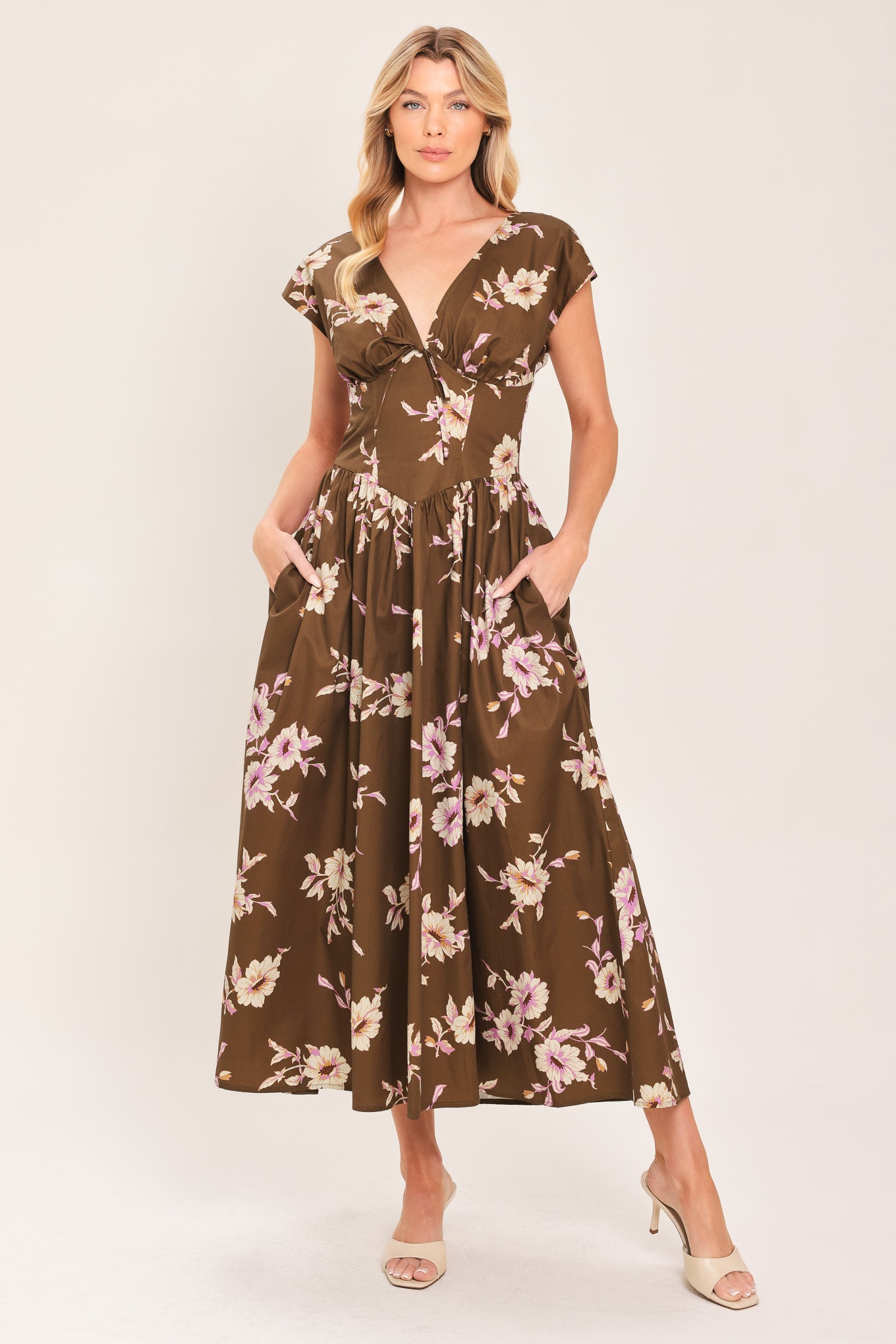 A woman wearing a brown midi dress with a pink and beige floral print, featuring cap sleeves, a deep V-neckline, and side pockets | Flying Tomato
