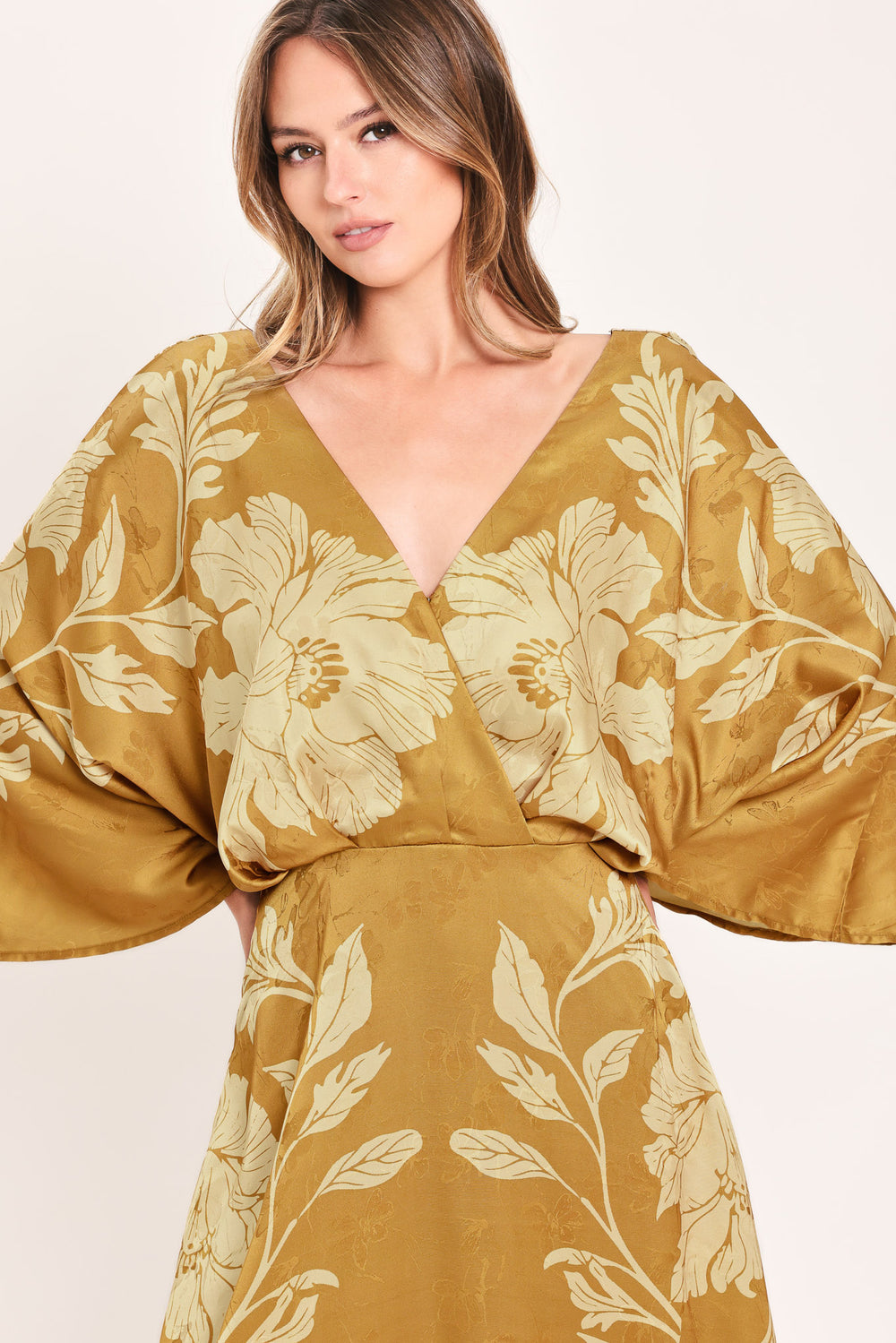 A woman wearing a midi-length, V-neck dress with wide bell sleeves in a golden camel fabric featuring a cream floral jacquard pattern | Flying Tomato