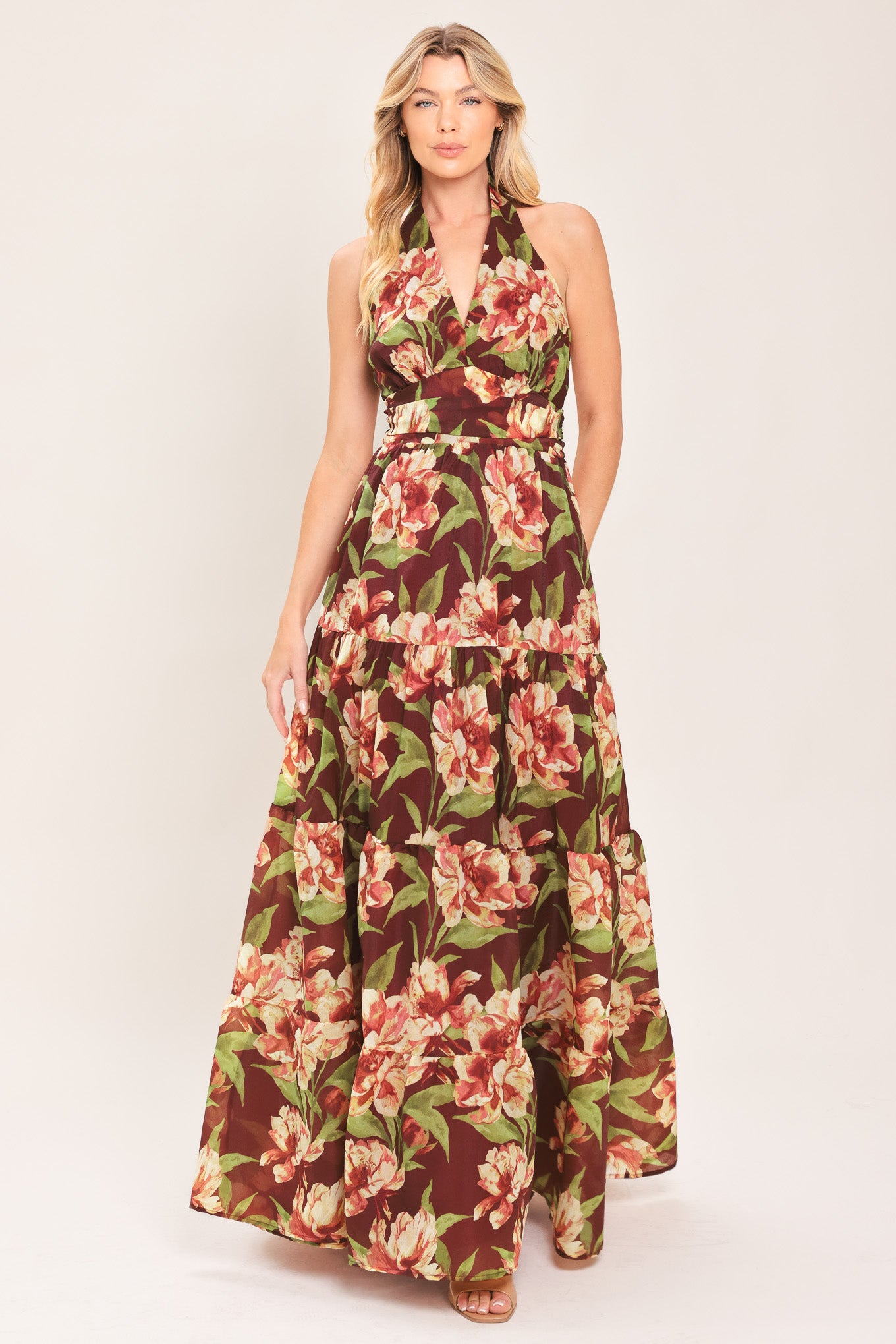 A woman wearing a halter-neck, tiered maxi dress with a burgundy base and a large pink and green floral print | Flying Tomato