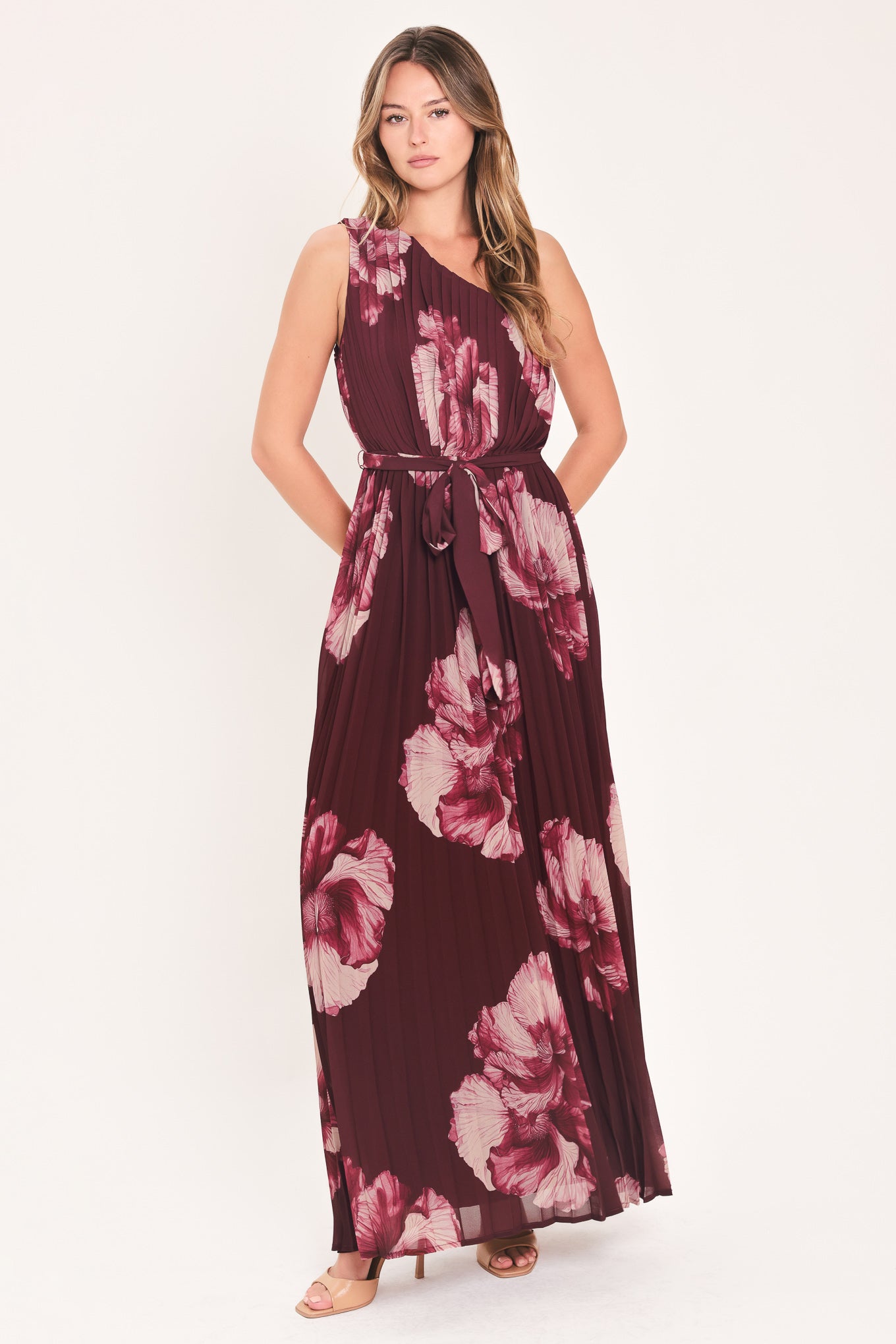 A woman modeling a sleeveless, one-shoulder maxi dress with a pleated skirt and waist tie. The dress features a dark burgundy base with a large floral print in dusty pink and mauve tones | Flying Tomato