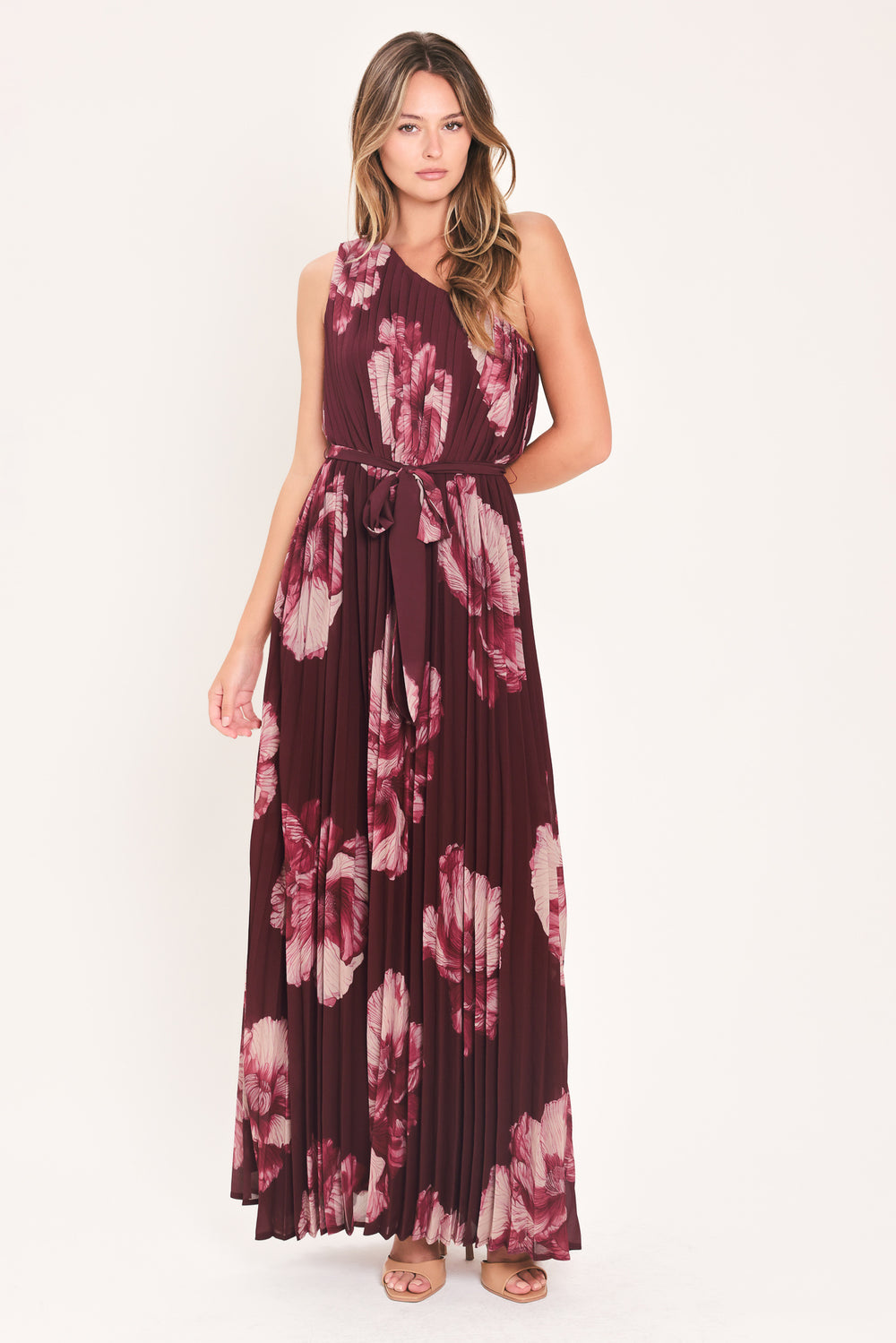 A woman modeling a sleeveless, one-shoulder maxi dress with a pleated skirt and waist tie. The dress features a dark burgundy base with a large floral print in dusty pink and mauve tones | Flying Tomato