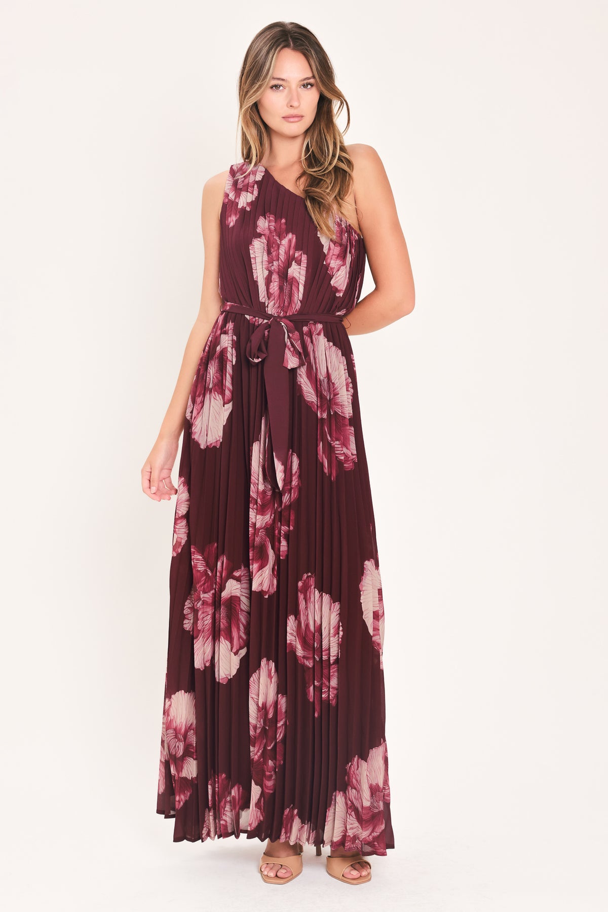 A woman modeling a sleeveless, one-shoulder maxi dress with a pleated skirt and waist tie. The dress features a dark burgundy base with a large floral print in dusty pink and mauve tones | Flying Tomato