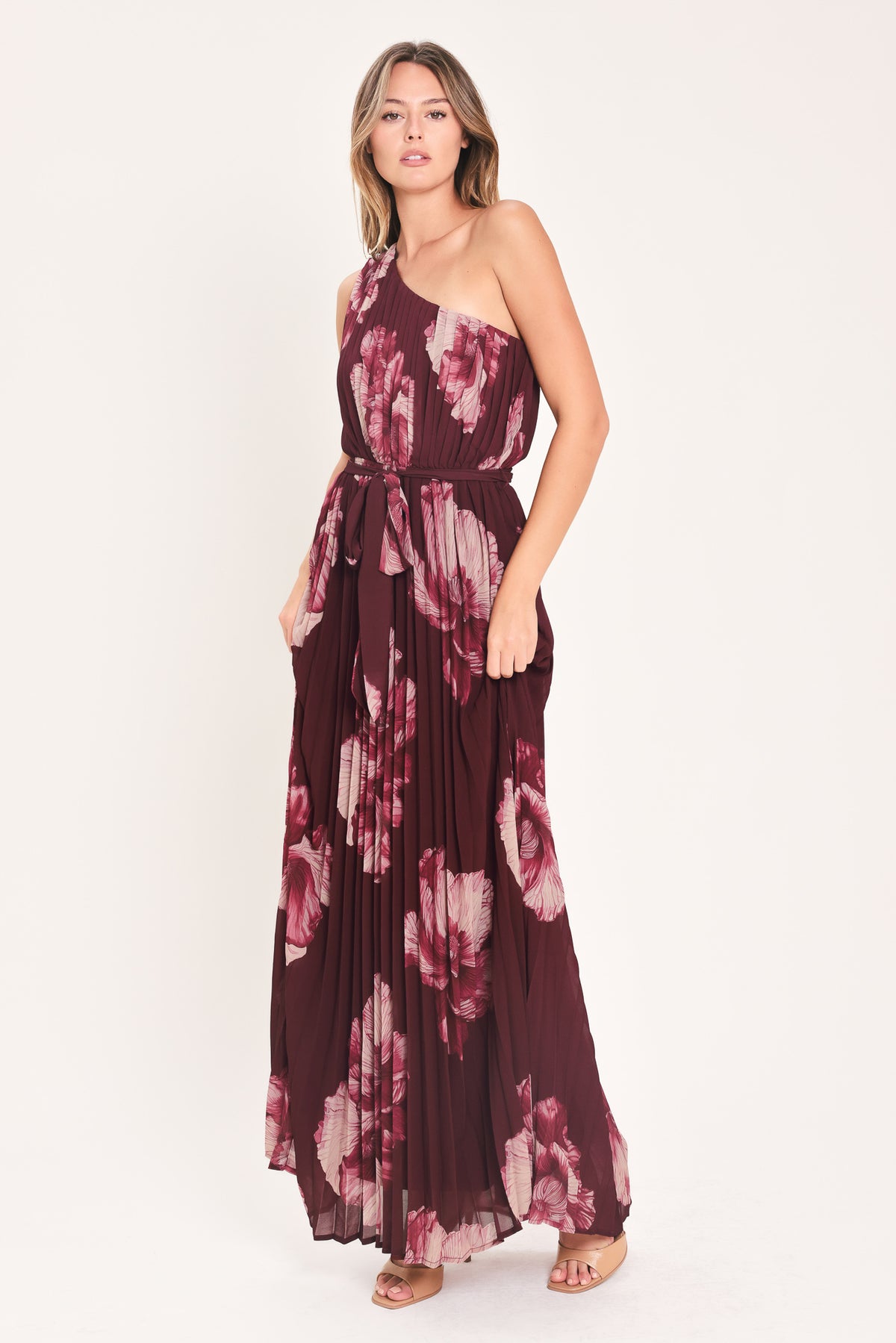 A woman modeling a sleeveless, one-shoulder maxi dress with a pleated skirt and waist tie. The dress features a dark burgundy base with a large floral print in dusty pink and mauve tones | Flying Tomato