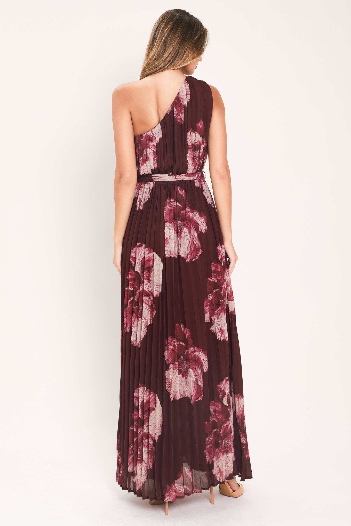A woman modeling a sleeveless, one-shoulder maxi dress with a pleated skirt and waist tie. The dress features a dark burgundy base with a large floral print in dusty pink and mauve tones | Flying Tomato