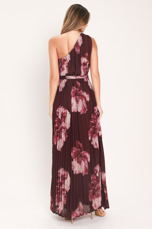 A woman modeling a sleeveless, one-shoulder maxi dress with a pleated skirt and waist tie. The dress features a dark burgundy base with a large floral print in dusty pink and mauve tones | Flying Tomato