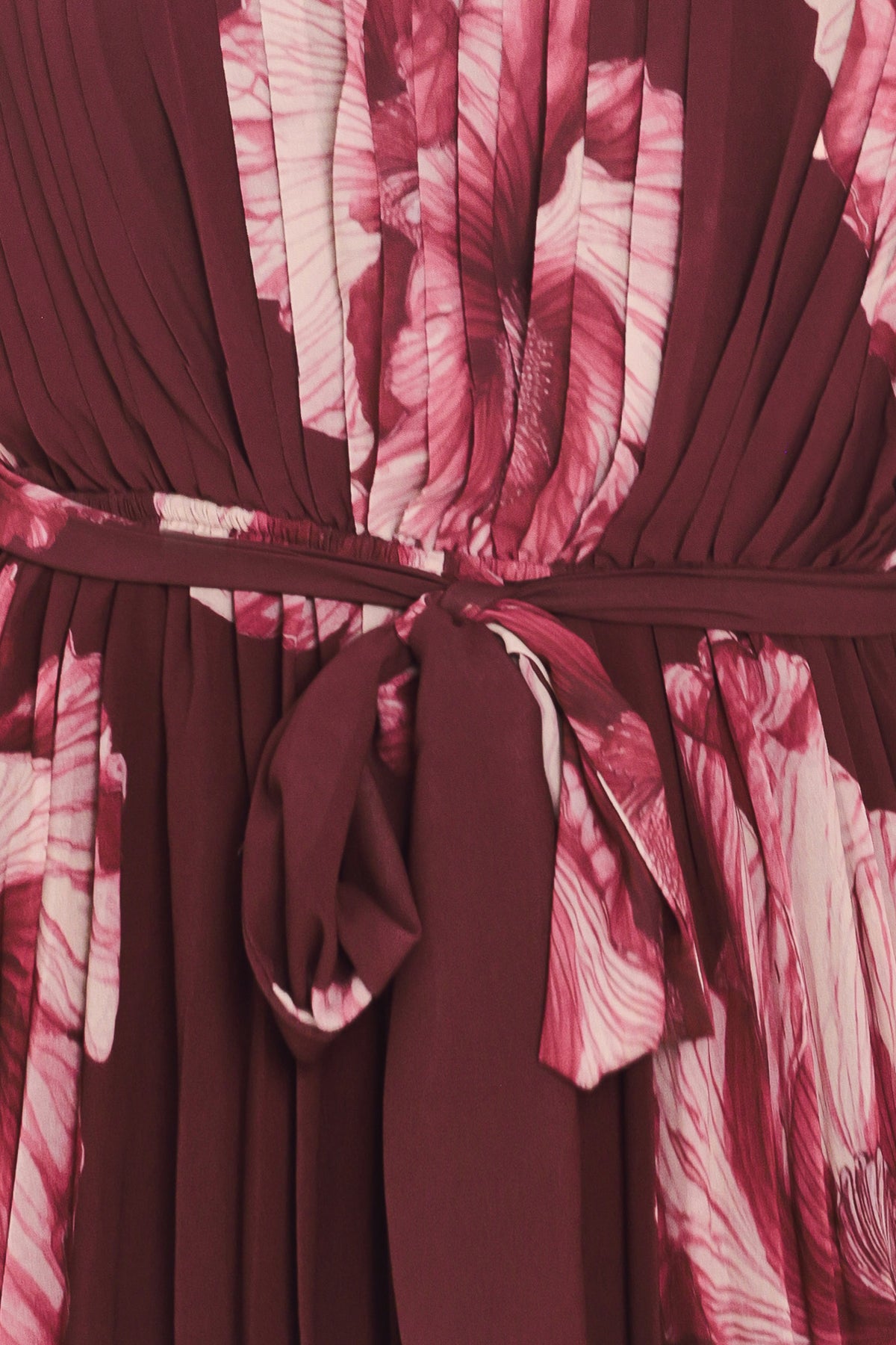 A woman modeling a sleeveless, one-shoulder maxi dress with a pleated skirt and waist tie. The dress features a dark burgundy base with a large floral print in dusty pink and mauve tones | Flying Tomato