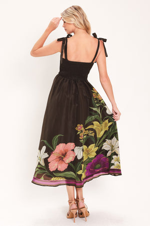 A woman models a sleeveless midi dress with a sweetheart neckline, shoulder straps, and a dark brown base featuring a colorful floral print along the hem and bodice | Flying Tomato