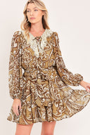 A woman wearing a long-sleeve camel mini dress with a brown and beige paisley pattern, featuring a white lace ruffle collar and a matching fabric tie belt at the waist | Flying Tomato