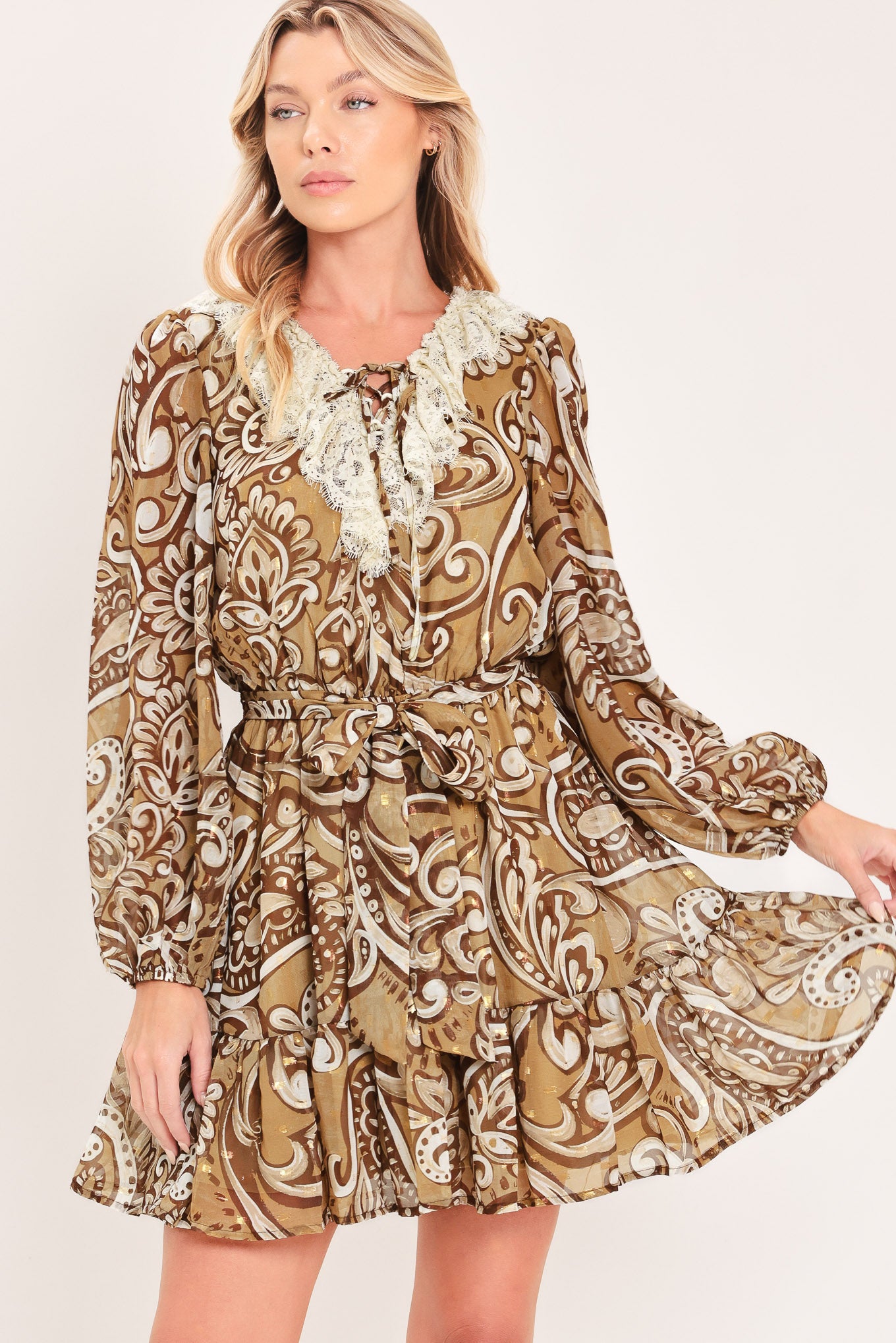 A woman wearing a long-sleeve camel mini dress with a brown and beige paisley pattern, featuring a white lace ruffle collar and a matching fabric tie belt at the waist | Flying Tomato