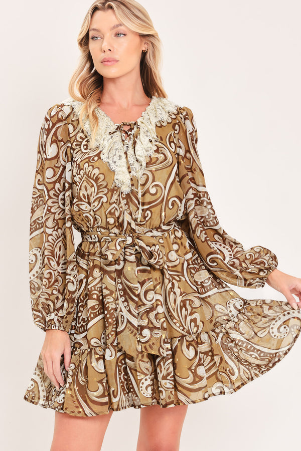 A woman wearing a long-sleeve camel mini dress with a brown and beige paisley pattern, featuring a white lace ruffle collar and a matching fabric tie belt at the waist | Flying Tomato
