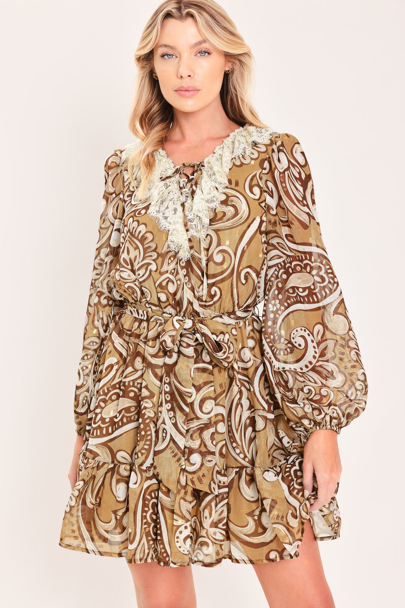 A woman wearing a long-sleeve camel mini dress with a brown and beige paisley pattern, featuring a white lace ruffle collar and a matching fabric tie belt at the waist | Flying Tomato