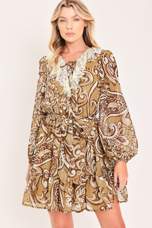 A woman wearing a long-sleeve camel mini dress with a brown and beige paisley pattern, featuring a white lace ruffle collar and a matching fabric tie belt at the waist | Flying Tomato
