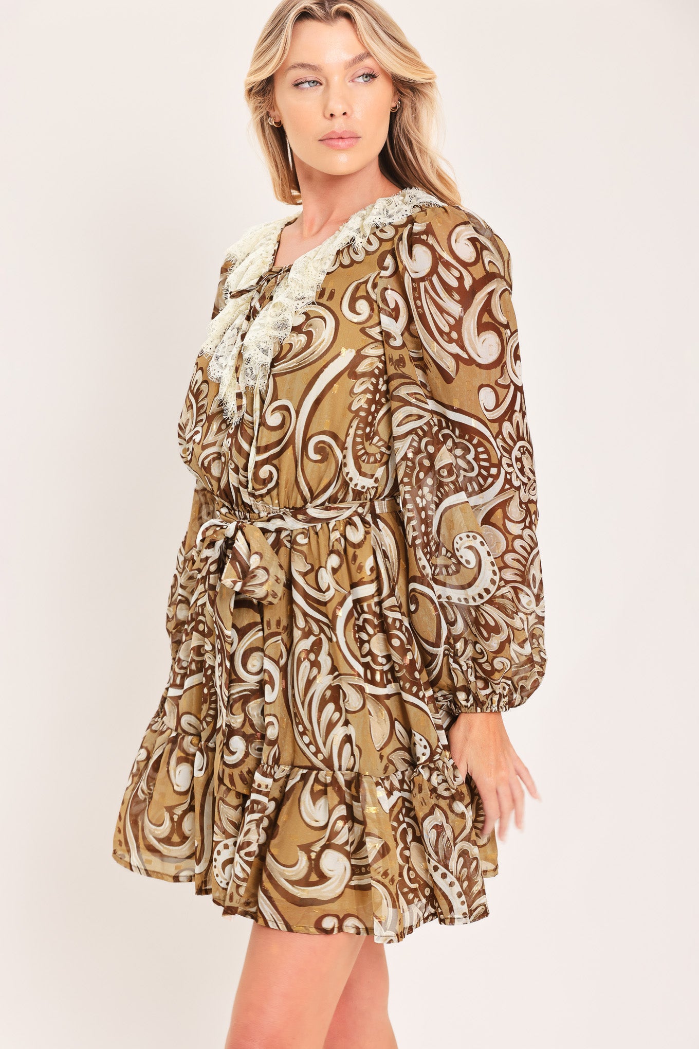 A woman wearing a long-sleeve camel mini dress with a brown and beige paisley pattern, featuring a white lace ruffle collar and a matching fabric tie belt at the waist | Flying Tomato