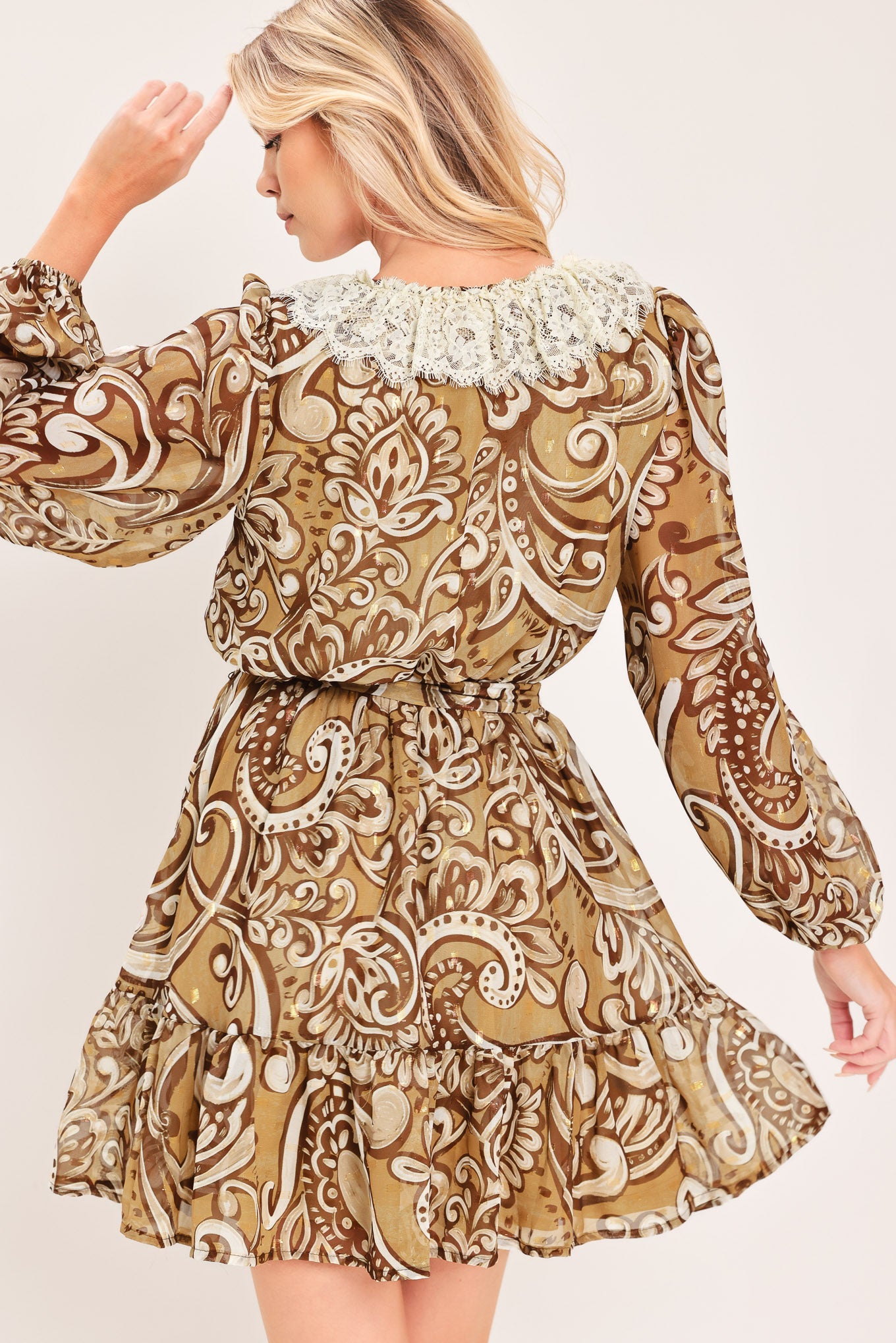 A woman wearing a long-sleeve camel mini dress with a brown and beige paisley pattern, featuring a white lace ruffle collar and a matching fabric tie belt at the waist | Flying Tomato