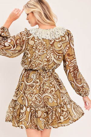 A woman wearing a long-sleeve camel mini dress with a brown and beige paisley pattern, featuring a white lace ruffle collar and a matching fabric tie belt at the waist | Flying Tomato