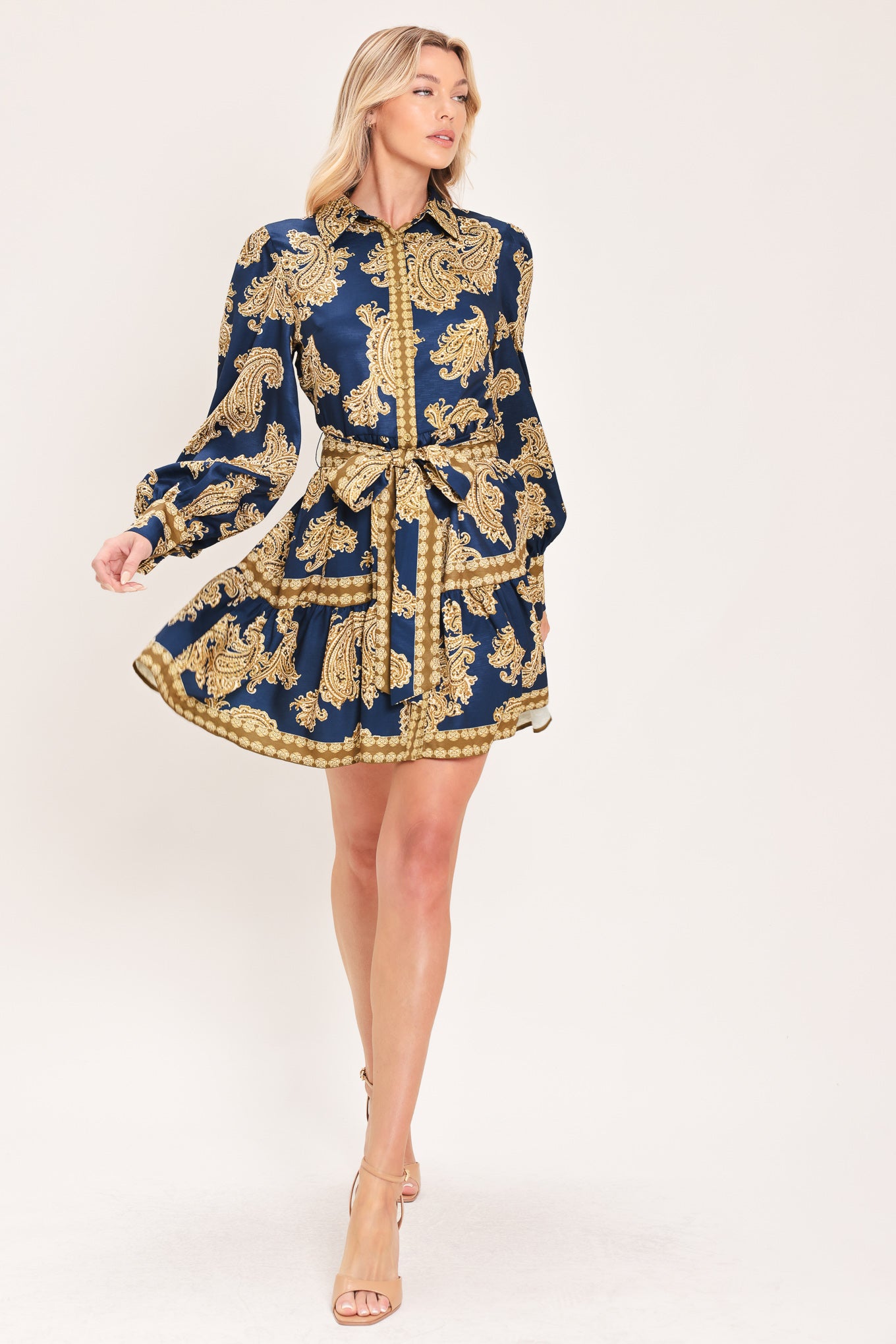 A woman modeling a long-sleeve mini dress with a blue and gold paisley pattern, featuring a button-up front, tie waist, and flared skirt | Flying Tomato