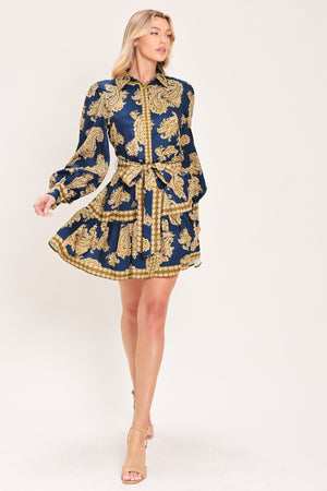 A woman modeling a long-sleeve mini dress with a blue and gold paisley pattern, featuring a button-up front, tie waist, and flared skirt | Flying Tomato