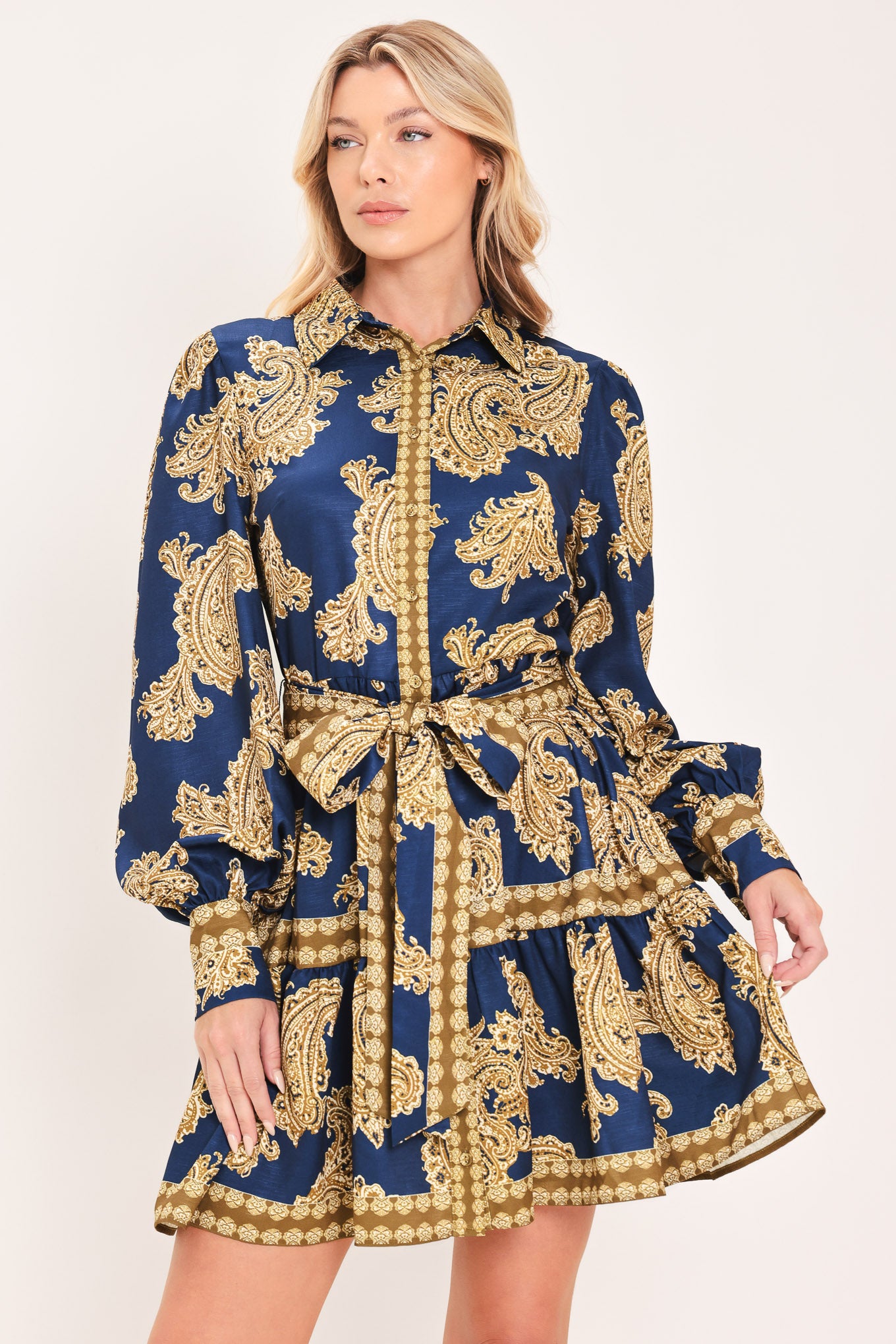 A woman modeling a long-sleeve mini dress with a blue and gold paisley pattern, featuring a button-up front, tie waist, and flared skirt | Flying Tomato