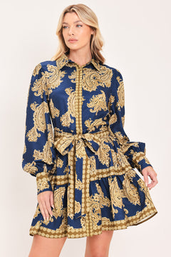 A woman modeling a long-sleeve mini dress with a blue and gold paisley pattern, featuring a button-up front, tie waist, and flared skirt | Flying Tomato