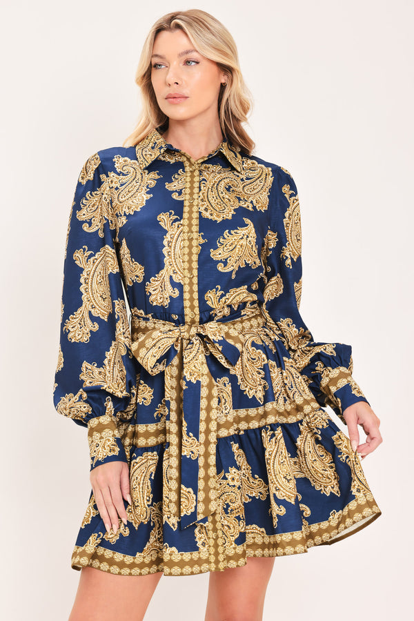 A woman modeling a long-sleeve mini dress with a blue and gold paisley pattern, featuring a button-up front, tie waist, and flared skirt | Flying Tomato