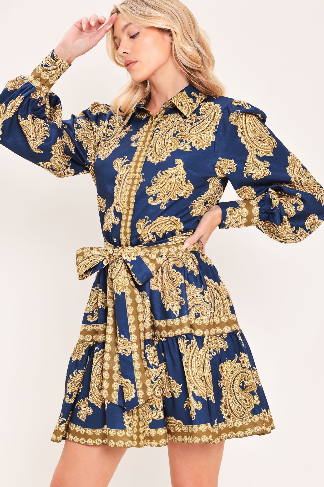 A woman modeling a long-sleeve mini dress with a blue and gold paisley pattern, featuring a button-up front, tie waist, and flared skirt | Flying Tomato