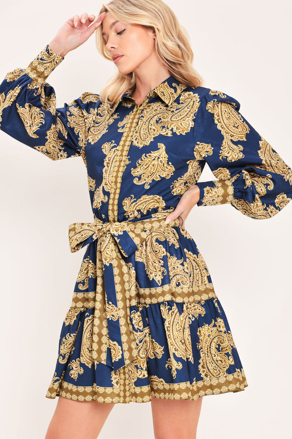 A woman modeling a long-sleeve mini dress with a blue and gold paisley pattern, featuring a button-up front, tie waist, and flared skirt | Flying Tomato