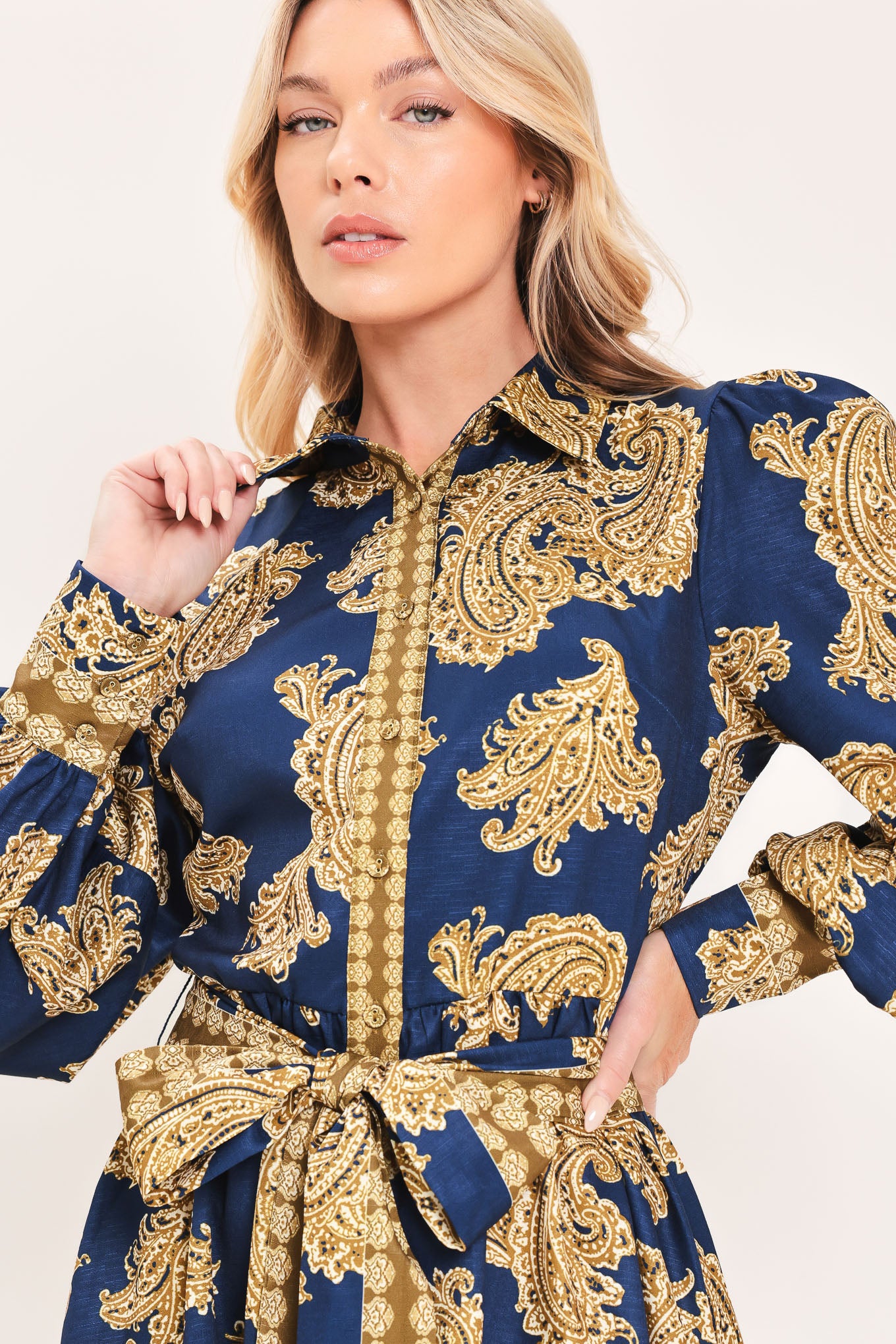A woman modeling a long-sleeve mini dress with a blue and gold paisley pattern, featuring a button-up front, tie waist, and flared skirt | Flying Tomato