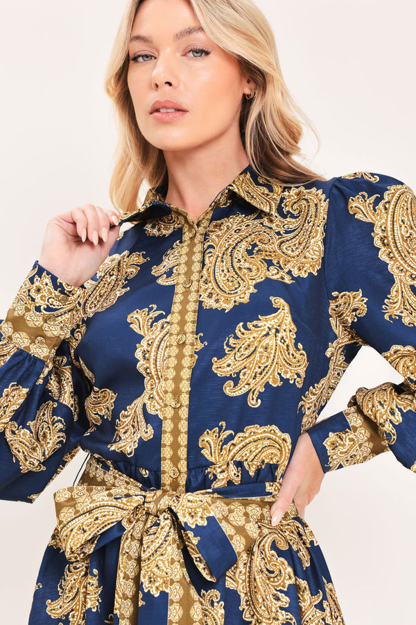 A woman modeling a long-sleeve mini dress with a blue and gold paisley pattern, featuring a button-up front, tie waist, and flared skirt | Flying Tomato