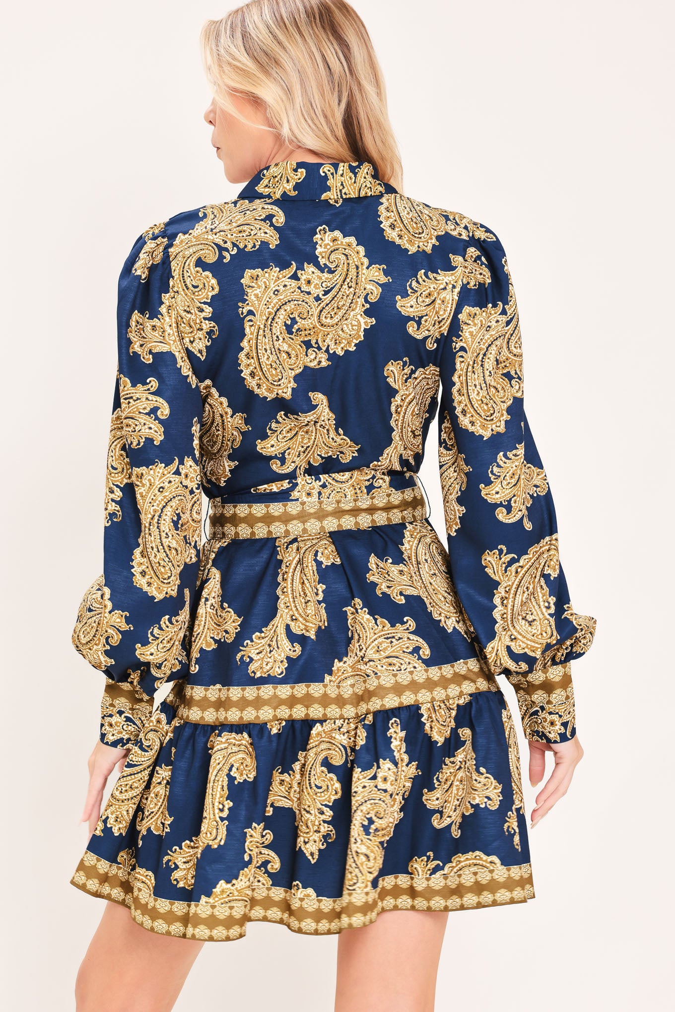 A woman modeling a long-sleeve mini dress with a blue and gold paisley pattern, featuring a button-up front, tie waist, and flared skirt | Flying Tomato