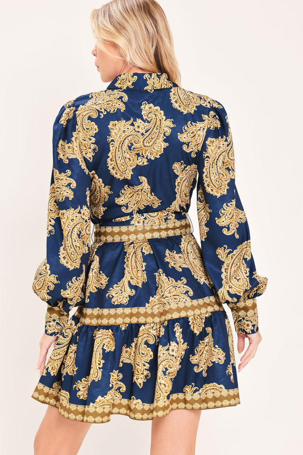 A woman modeling a long-sleeve mini dress with a blue and gold paisley pattern, featuring a button-up front, tie waist, and flared skirt | Flying Tomato